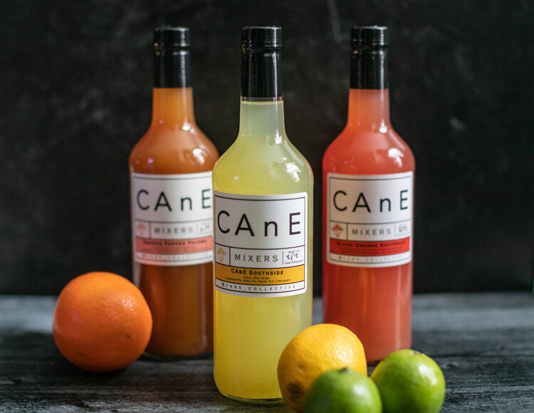 three bottles of CAnE Mixers on a table with an orange, lemon and limes